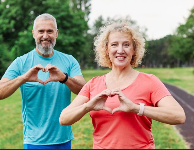 Kuvan etualalla keski-ikäinen nainen ja takana ikääntynyt mies, jotka molemmat muodostavat käsillään sydämen kuvan. Taustalla vihreää puistoa, kävelytie ja lehtipuita.